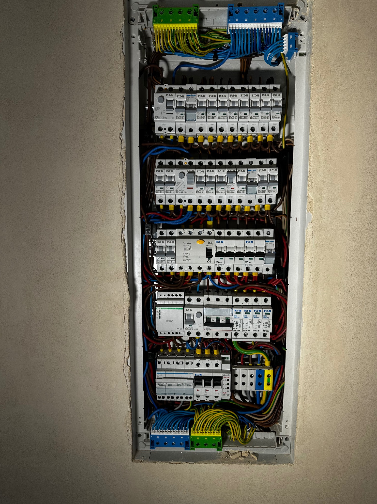 Rozdzielnica elektryczna z zabezpieczeniami nadprądowymi i różnicowoprądowymi, widoczne przewody w różnych kolorach, zamontowana w ścianie z widocznymi śladami po montażu.