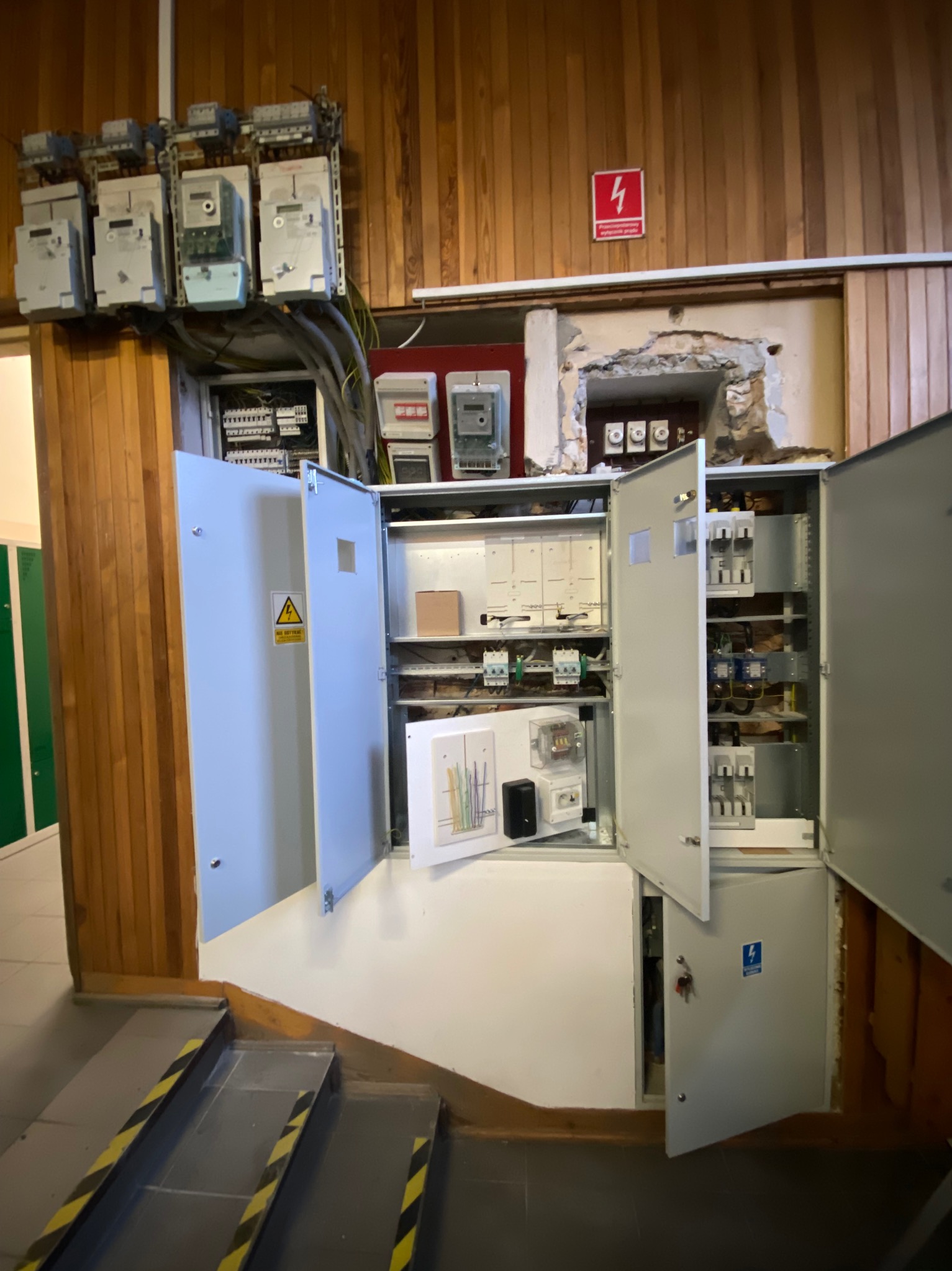 Otwarta szafa elektryczna z licznikami na drewnianej ścianie, widoczne przewody i zabezpieczenia, obok schody z żółto-czarnymi pasami ostrzegawczymi.