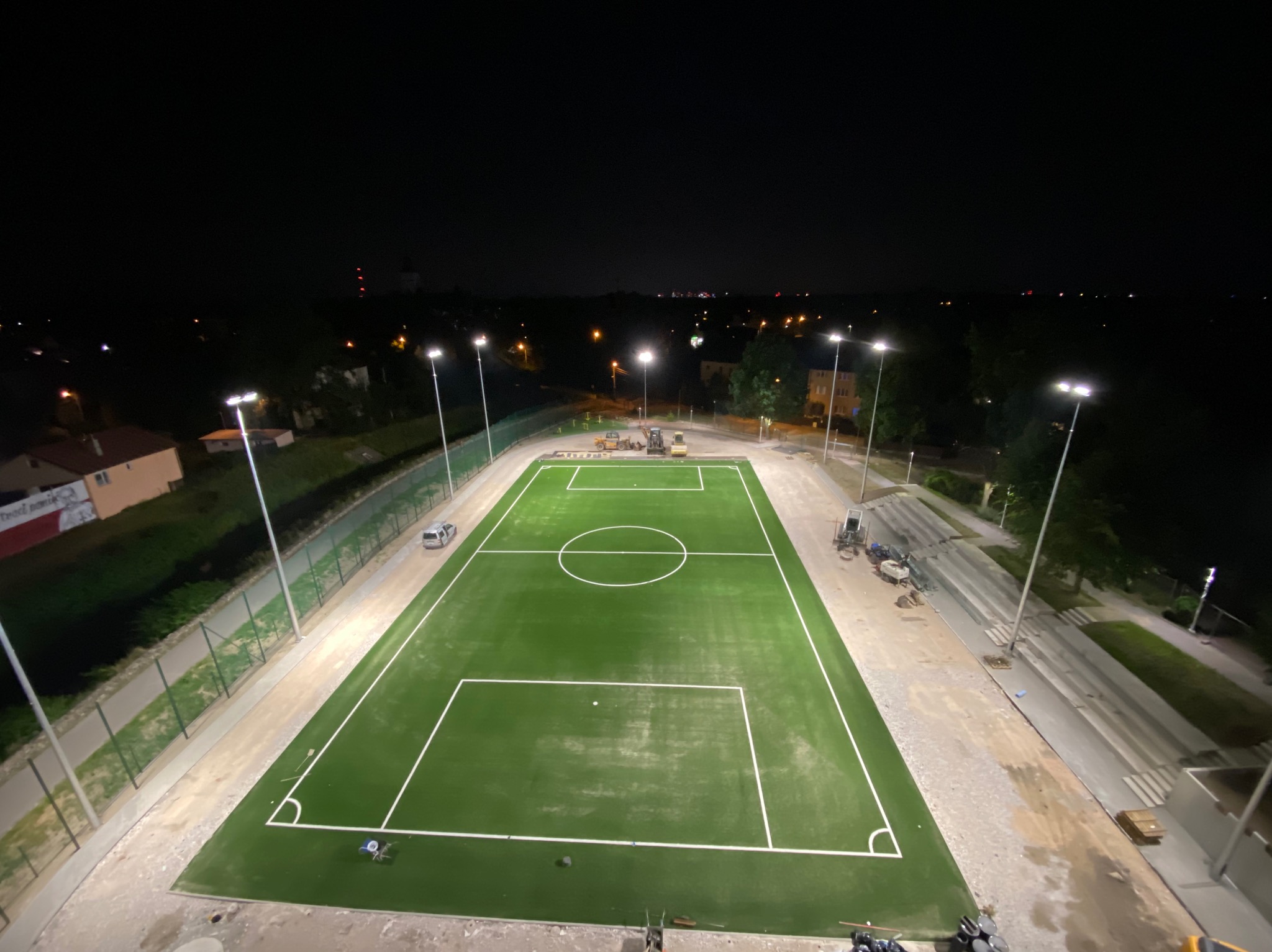Oświetlony nocą, widok z góry na nowo wybudowane boisko z zieloną sztuczną nawierzchnią, białe linie boiska i słupy oświetleniowe, w tle zabudowania miejskie i maszyny budowlane.