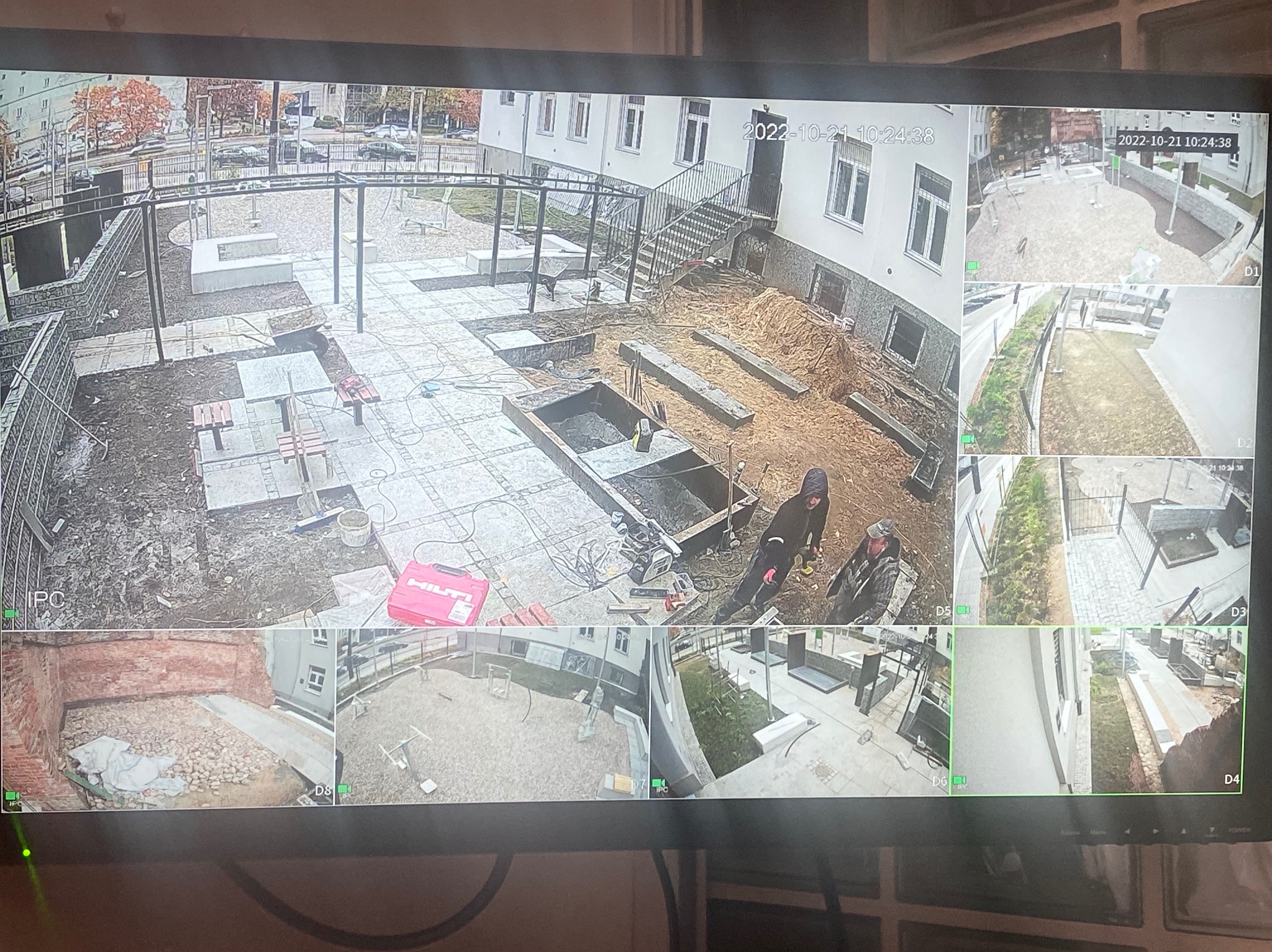 Monitoring placu budowy z widokiem na prace ziemne, układanie kostki brukowej i budowę elementów małej architektury, widoczne osoby wykonujące prace. Data i godzina na ekranie monitoringu.