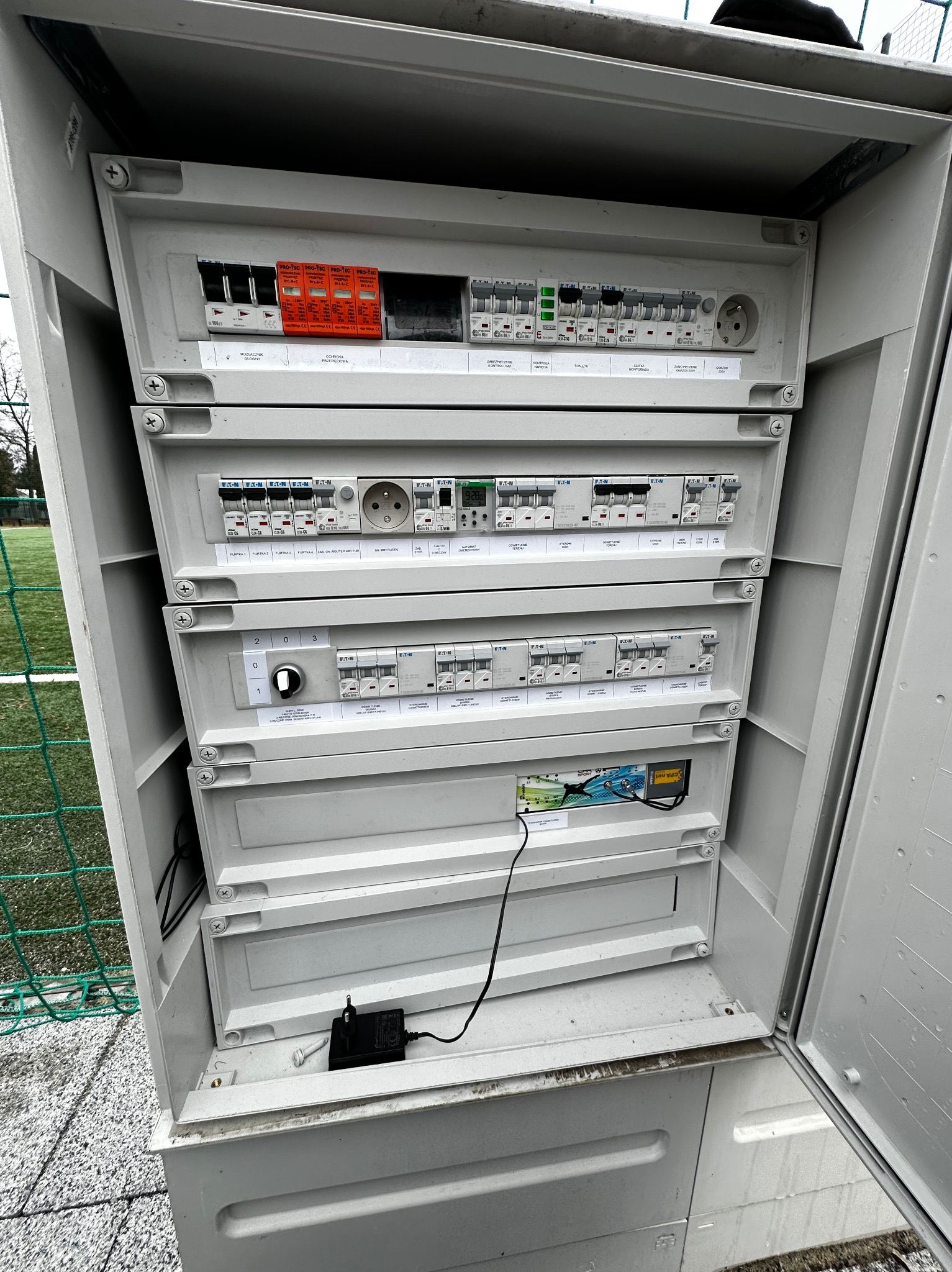 Otwarta szafa elektryczna z widocznymi bezpiecznikami, wyłącznikami i gniazdkami, umieszczona na zewnątrz budynku, z fragmentem trawnika i ogrodzenia w tle.