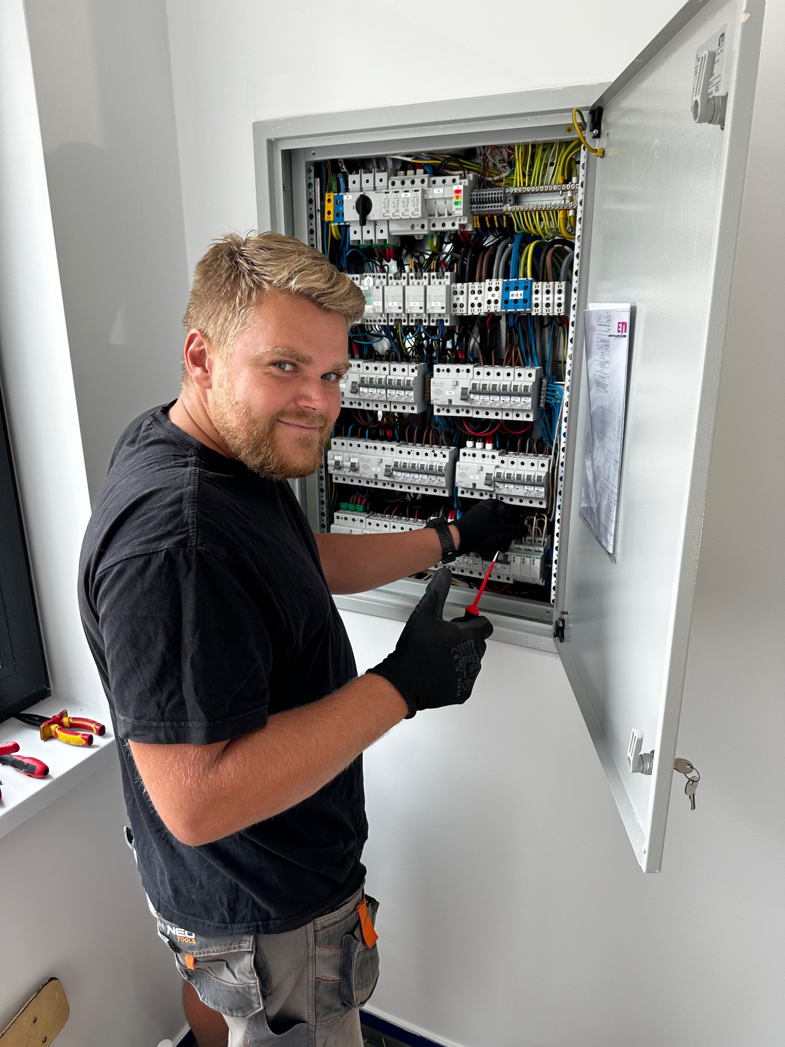 Uśmiechnięty elektryk w czarnych rękawicach, używający czerwonego śrubokrętu do pracy w rozdzielnicy elektrycznej z otwartymi drzwiami, widoczny schemat na wewnętrznej stronie drzwi