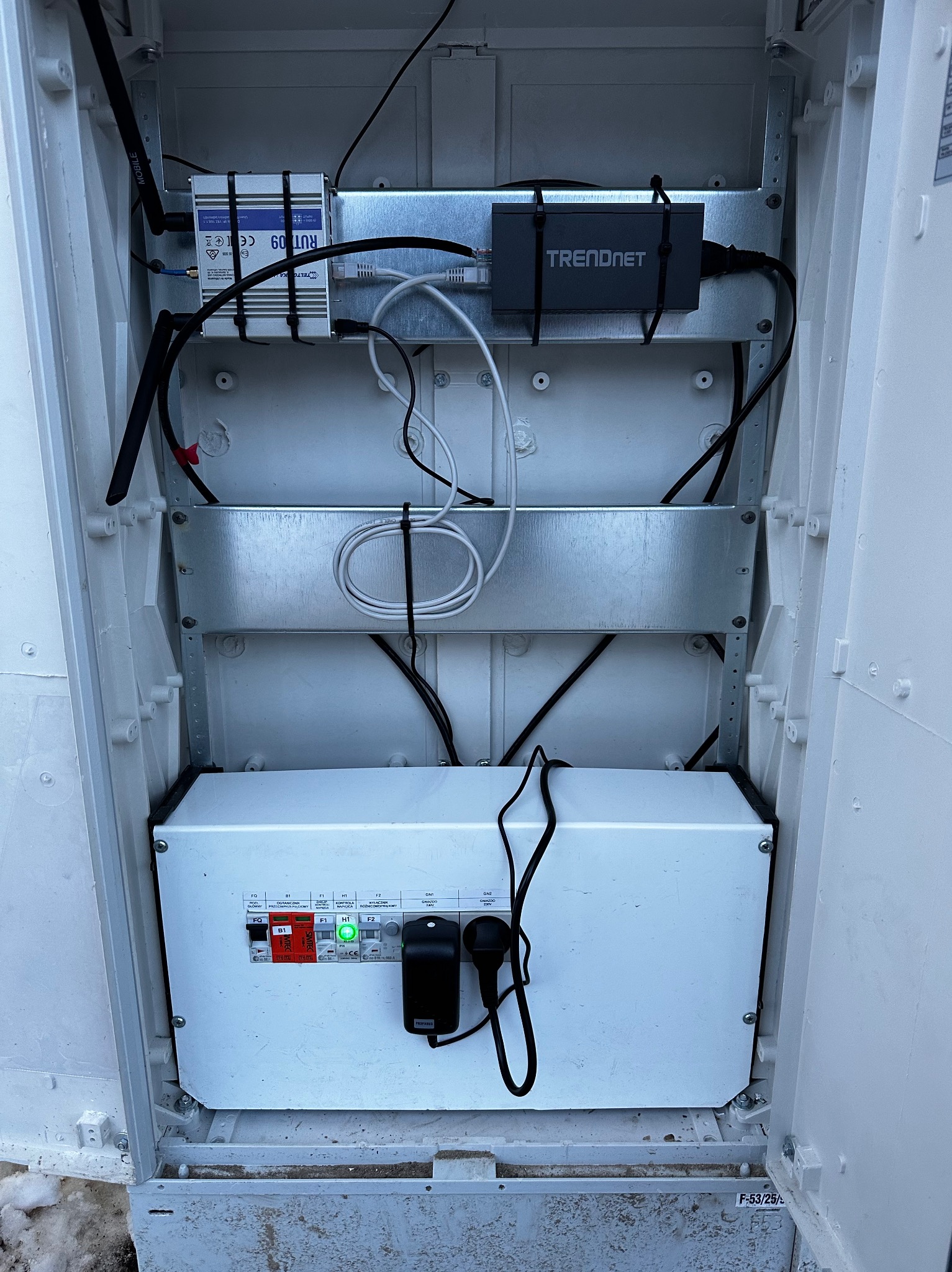 Otwarta szafka telekomunikacyjna z zamontowanymi urządzeniami sieciowymi: router RUT 09, urządzenie Trendnet, okablowanie i listwa zasilająca z włącznikiem i diodami LED.