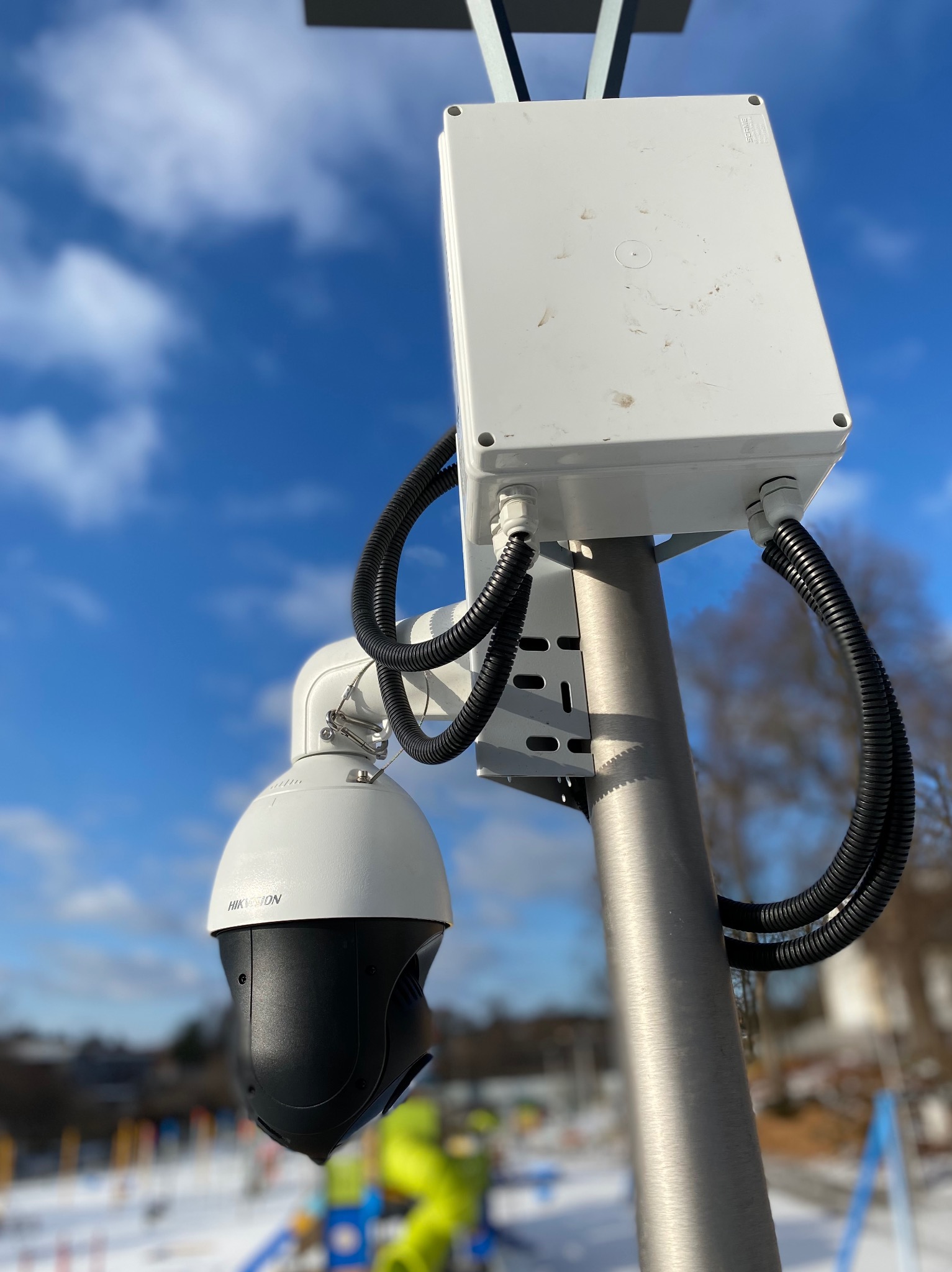 Kamera przemysłowa PTZ marki Hikvision zamontowana na metalowym słupie z białą puszką instalacyjną, widoczne przewody w peszlach, w tle błękitne niebo i plac zabaw.