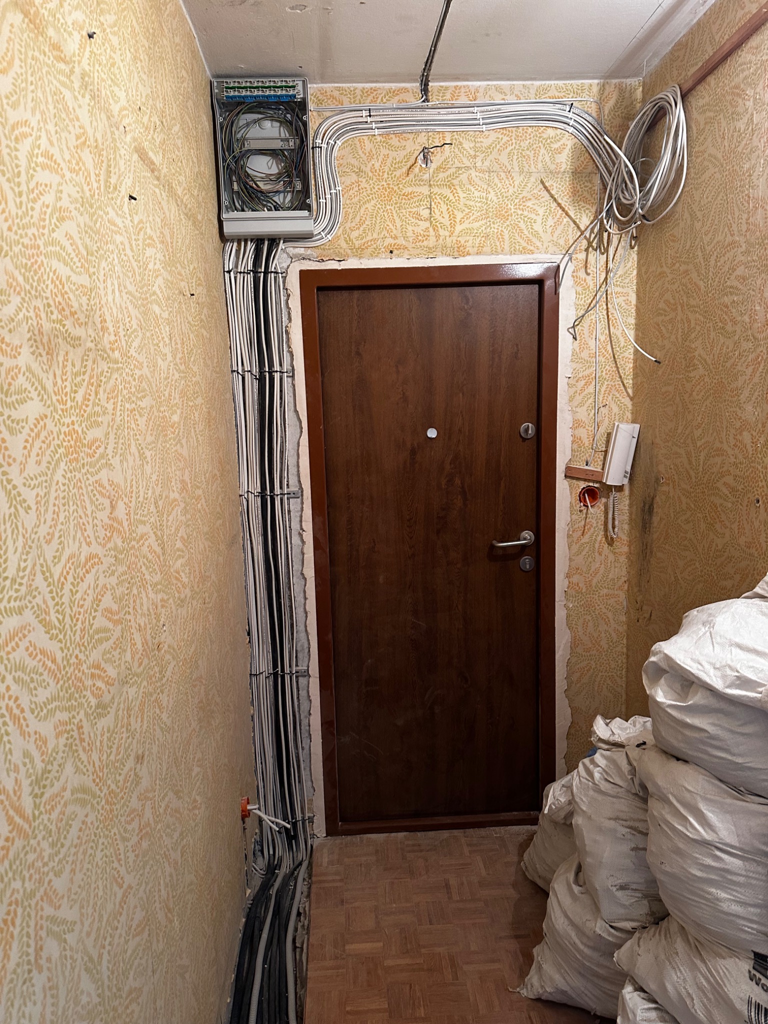 Wnętrze w trakcie remontu z widoczną skrzynką elektryczną, przewodami ułożonymi w korytkach na ścianie z wzorzystą tapetą, oraz stertą worków z materiałami budowlanymi przy brązowych drzwiach.
