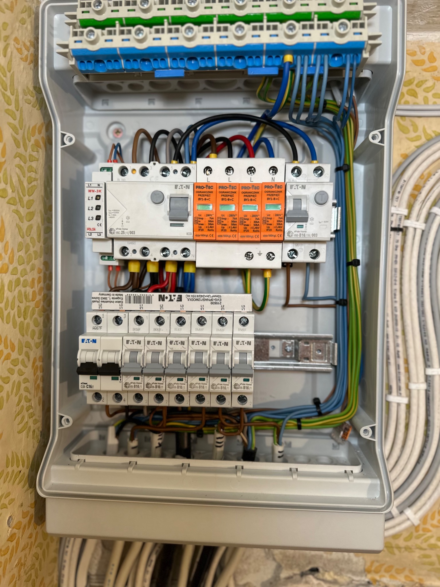 Otwarta skrzynka rozdzielcza z zainstalowanymi bezpiecznikami, ogranicznikami przepięć i okablowaniem, widoczne brązowe, żółte, niebieskie i zielone przewody, zasilanie trójfazowe.