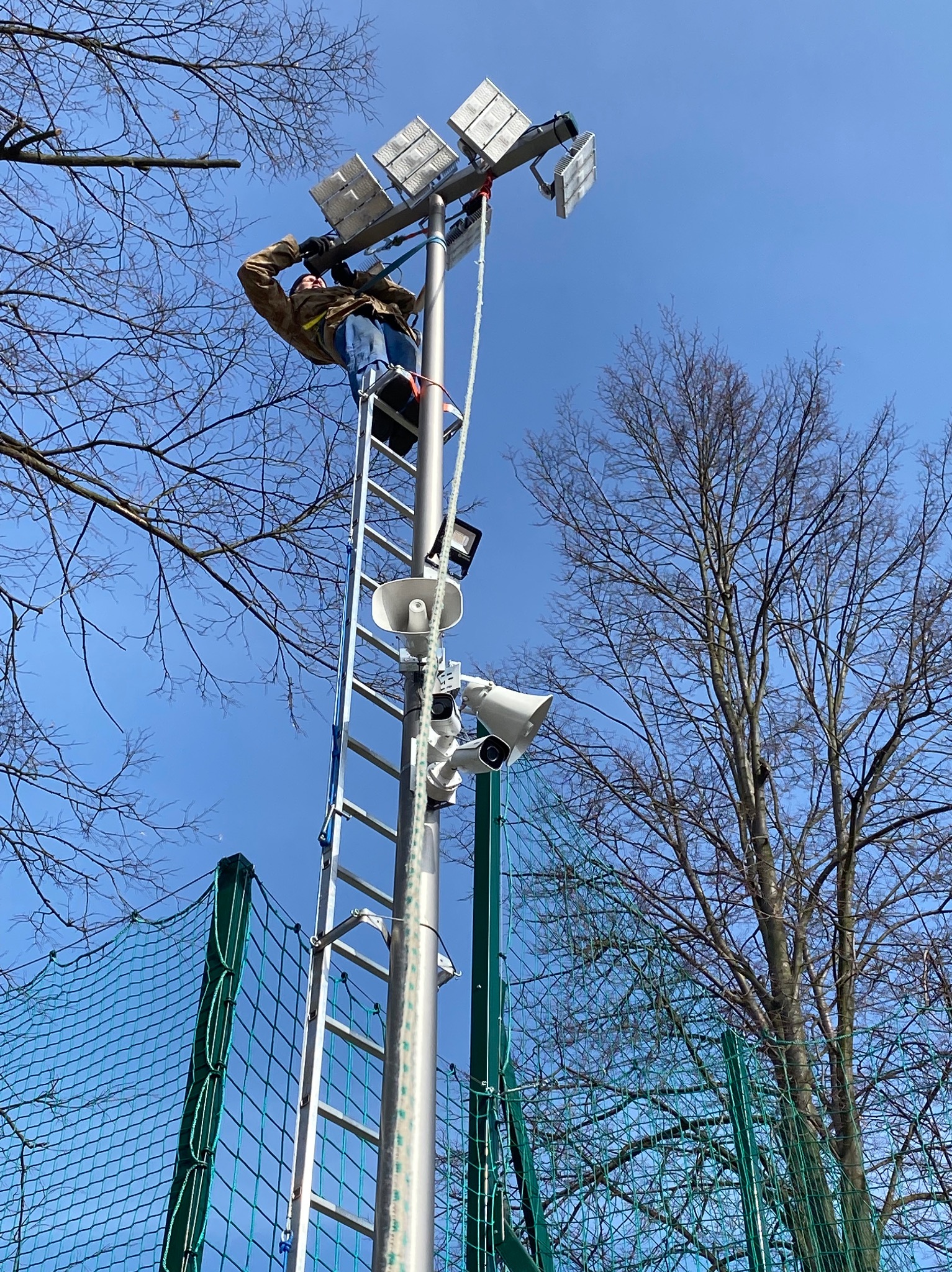 Pracownik na drabinie montuje lub serwisuje reflektory i głośniki na wysokim maszcie oświetleniowym, widoczne drzewa i siatka ogrodzeniowa w tle.