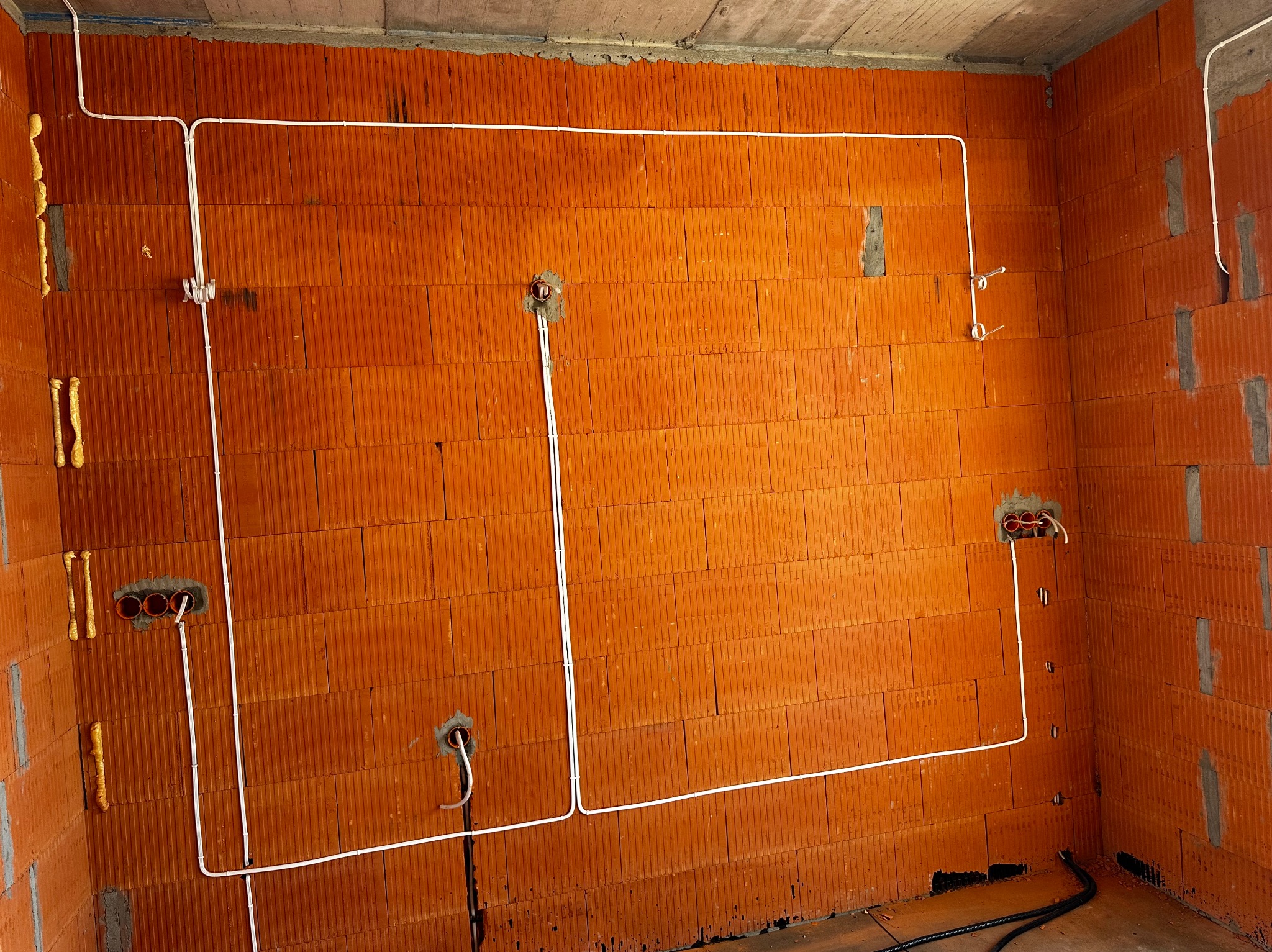 Instalacja elektryczna natynkowa w nowym budynku z czerwonej cegły dziurawki. Białe kable prowadzone wzdłuż ścian, widoczne puszki pod gniazdka i przewody.