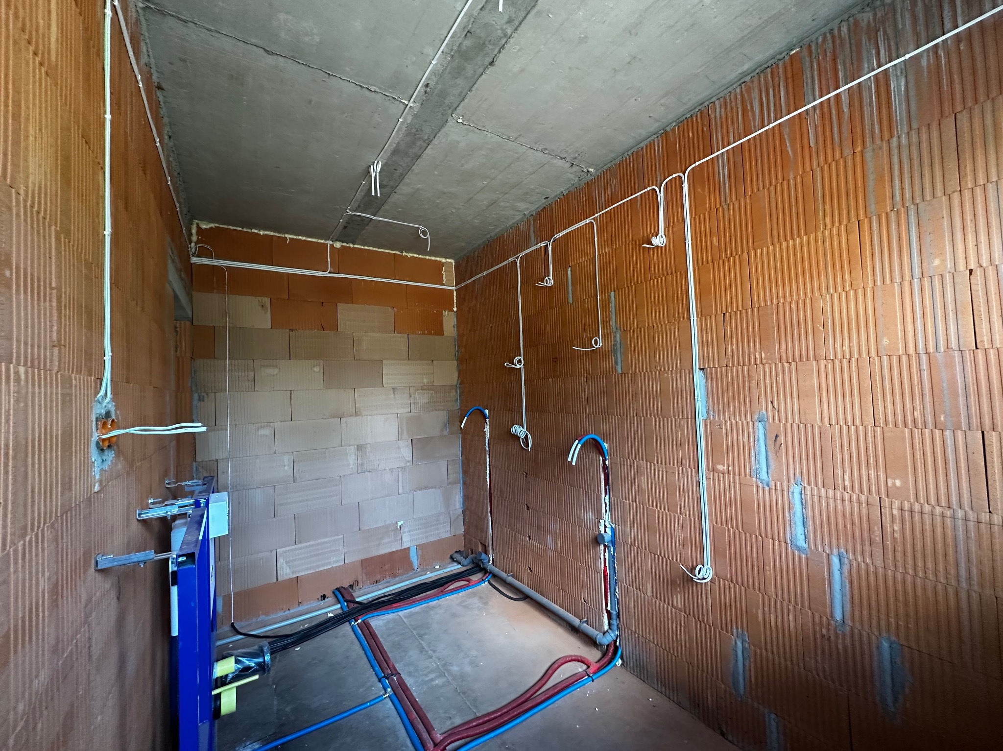 Surowy stan łazienki w bloku z widocznymi przewodami elektrycznymi i instalacją wodną w ścianach z czerwonej cegły i betonowym sufitem. Instalacje przed tynkowaniem.