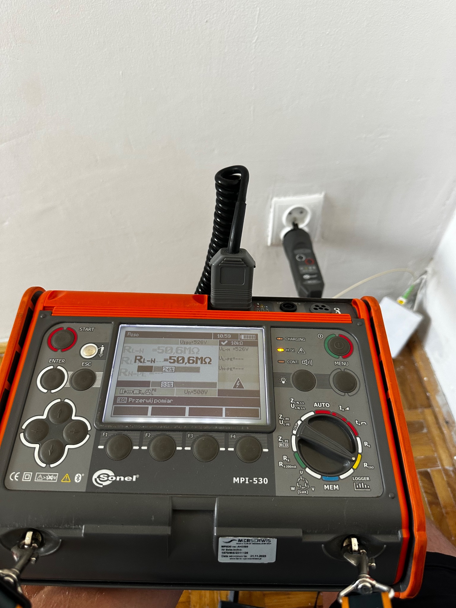 Miernik Sonel MPI-530 w trakcie pomiarów elektrycznych, widoczny ekran z wynikami, gniazdko z podłączonym przewodem pomiarowym. Urządzenie na tle ściany i podłogi.