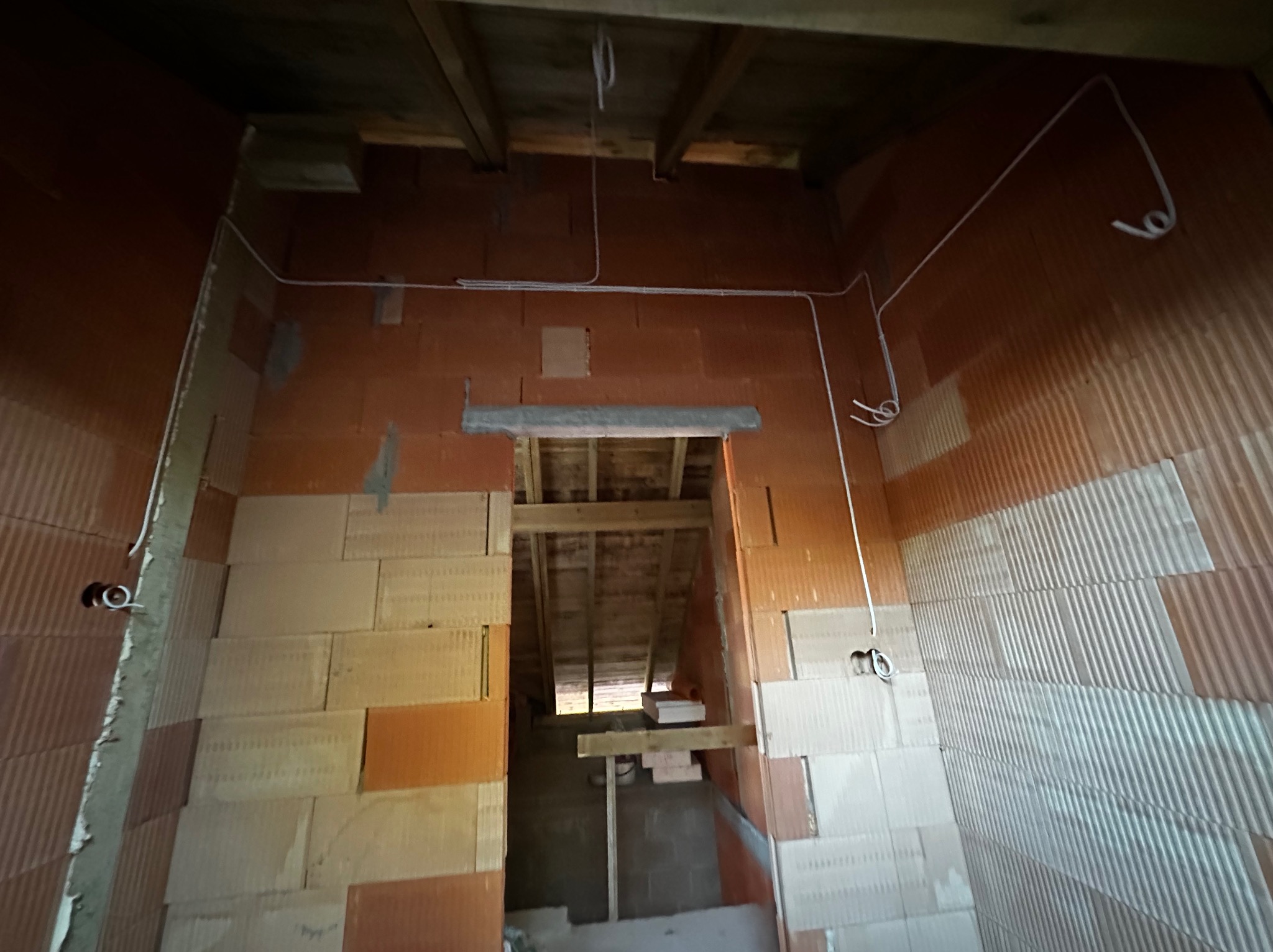 Wnętrze w trakcie budowy z widoczną instalacją elektryczną: przewody poprowadzone po ścianach z cegły, puszki elektryczne. Widok na surowy strop i przejście do kolejnego pomieszczenia.