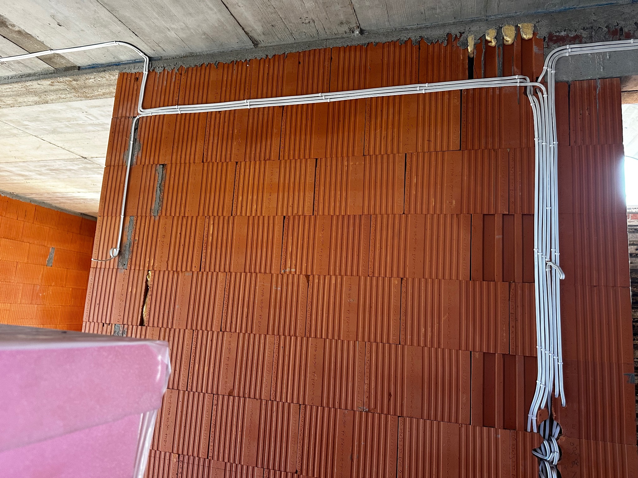 Białe przewody elektryczne ułożone na ścianie z czerwonej cegły dziurawki w surowym stanie. Widoczne mocowania przewodów. Instalacja w trakcie budowy.