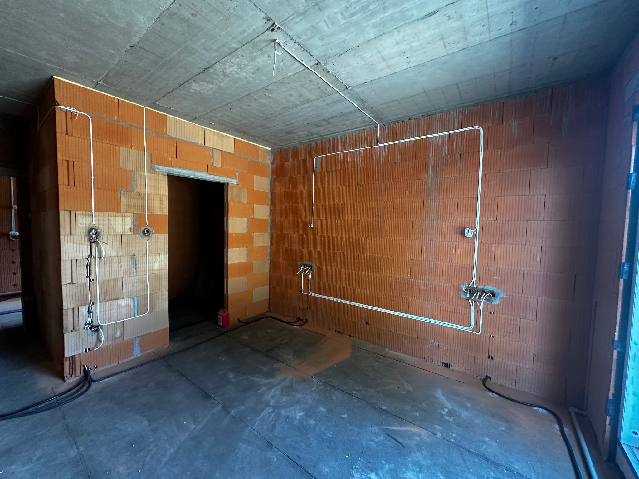 Surowy pokój z ceglanymi ścianami i betonowym sufitem. Widoczne białe kable elektryczne prowadzone po ścianach i suficie, przygotowane do montażu gniazdek i oświetlenia.