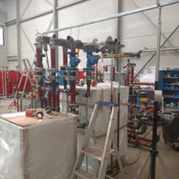 Rozbudowana instalacja rurowa z zaworami i elementami łączeniowymi w hali produkcyjnej, częściowo zabezpieczona folią, drabina oparta o obudowę.