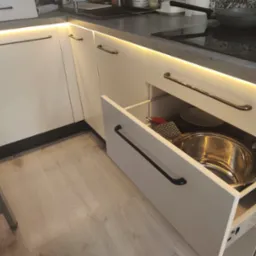 Otwarta szuflada w szafce kuchennej z białymi frontami i podświetleniem LED, widoczne garnki i naczynia wewnątrz, obok krzesło z motywem flagi brytyjskiej.