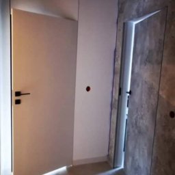 MGG spolka z o.o. - Białe, częściowo otwarte drzwi z czarną klamką, przylegające do białej ściany z widocznym gniazdkiem elektrycznym, obok drzwi we wnęce wykończonej szarą, betonową okładziną.