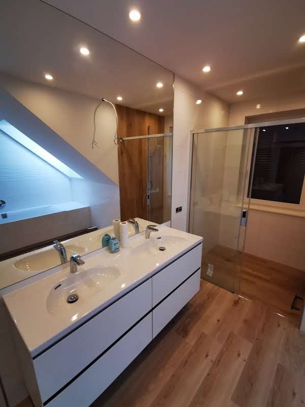 Nowoczesna łazienka na poddaszu z dwiema umywalkami w białej szafce, drewnianą podłogą i kabiną prysznicową w rogu, oświetlona punktowymi lampami.