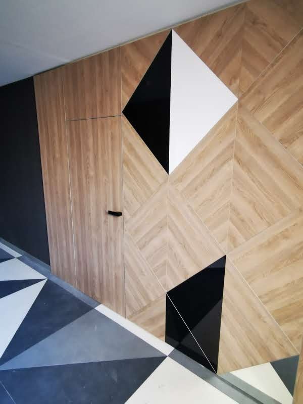 Ściana z drewnopodobnych płytek ułożonych w geometryczny wzór, uzupełniona czarnymi i białymi elementami w kształcie rombów, z ukrytymi drzwiami i podłogą w trójkątne wzory.