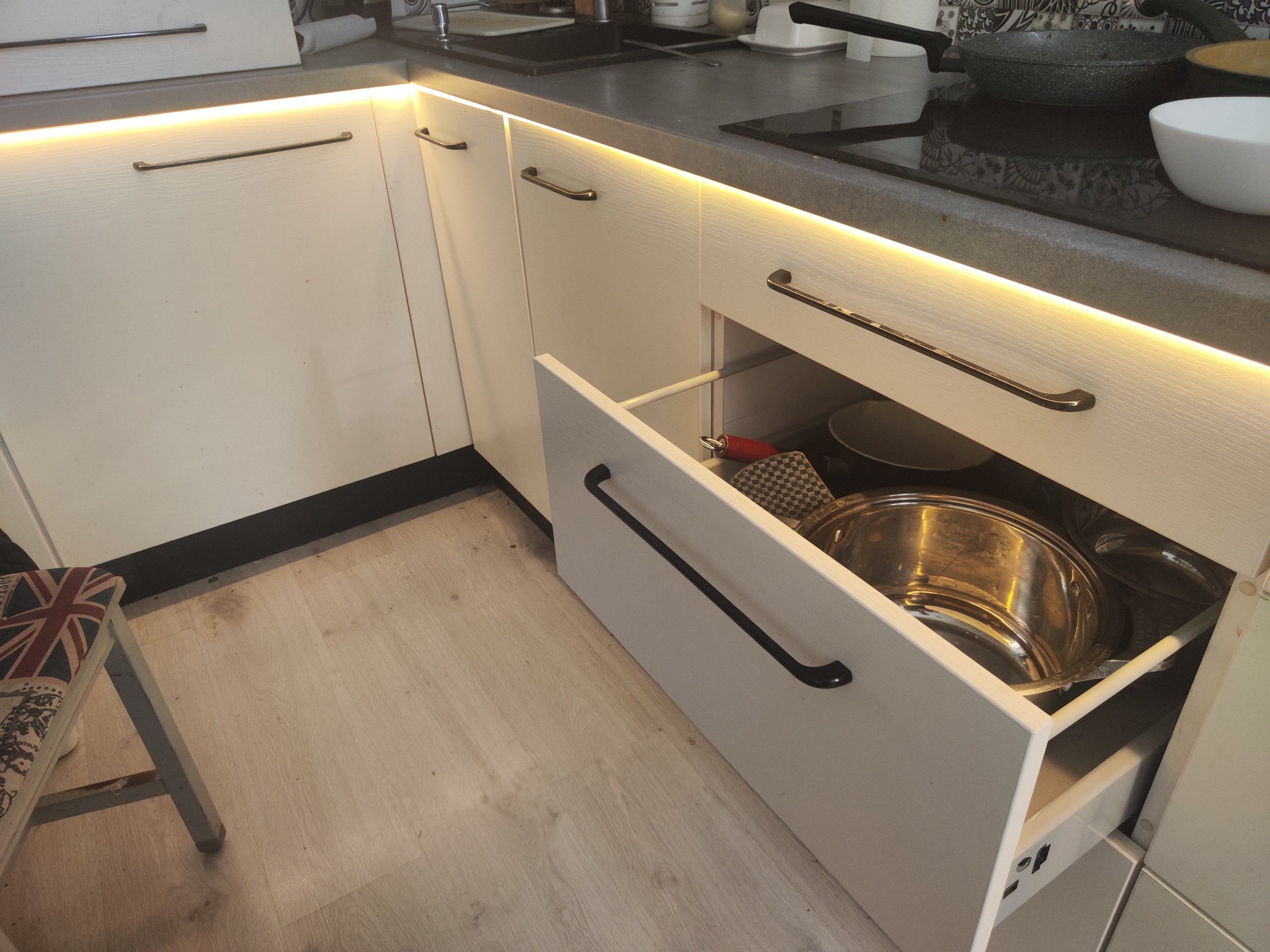 Otwarta szuflada w szafce kuchennej z białymi frontami i podświetleniem LED, widoczne garnki i naczynia wewnątrz, obok krzesło z motywem flagi brytyjskiej.