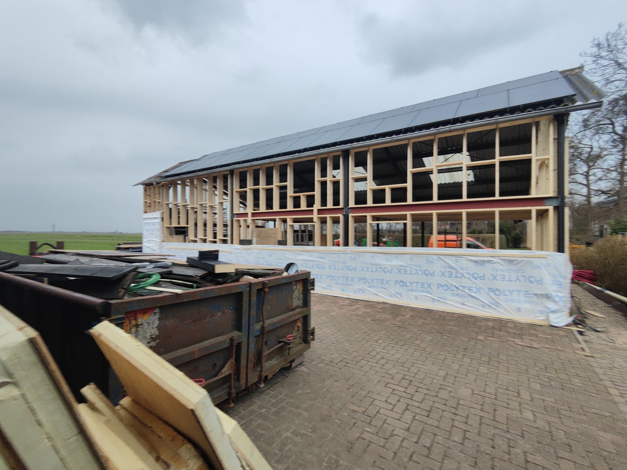 Konstrukcja szkieletowa budynku w trakcie budowy, widoczne drewniane ramy, stalowe belki, częściowo zamontowany dach i kontener ze śmieciami budowlanymi na pierwszym planie.