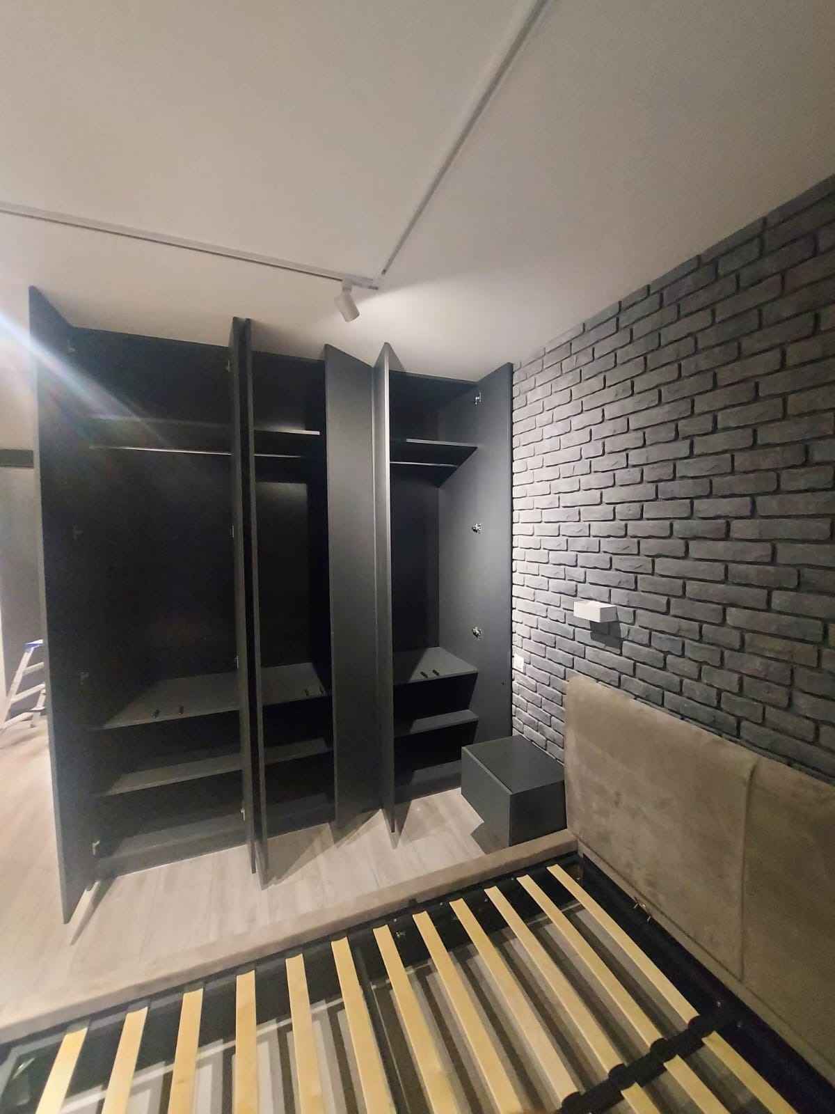 Nowoczesna szafa wnękowa z otwartymi drzwiami, w kolorze grafitowym, na tle ściany z szarej cegły dekoracyjnej, widoczny fragment ramy łóżka.