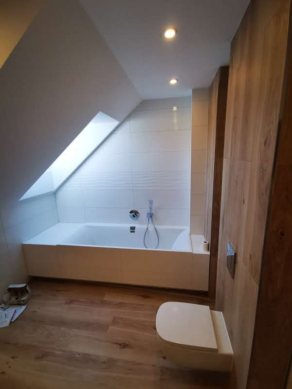 Nowoczesna łazienka na poddaszu z drewnianą podłogą i akcentami, biała wanna pod oknem dachowym, wisząca toaleta, oświetlenie punktowe.