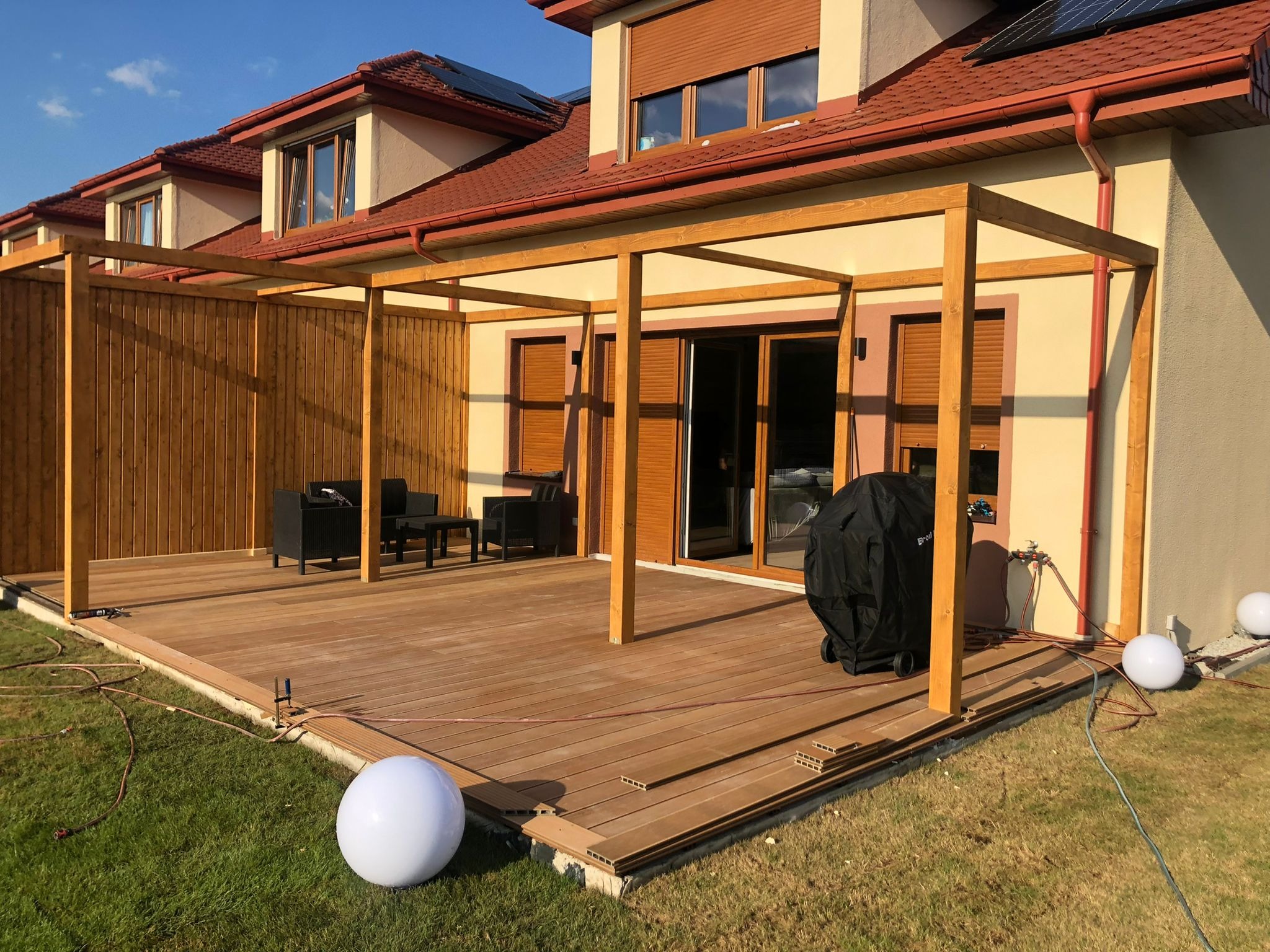 Nowoczesny taras z desek kompozytowych w trakcie budowy, widoczna konstrukcja z drewna, meble ogrodowe i grill, elementy oświetlenia w postaci kul.