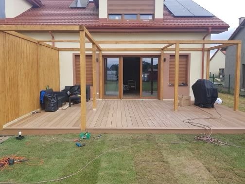 Montaż drewnianego tarasu z konstrukcją pergoli w trakcie realizacji, widoczne narzędzia na trawie, częściowo wyposażony meblami ogrodowymi, dom jednorodzinny w tle.