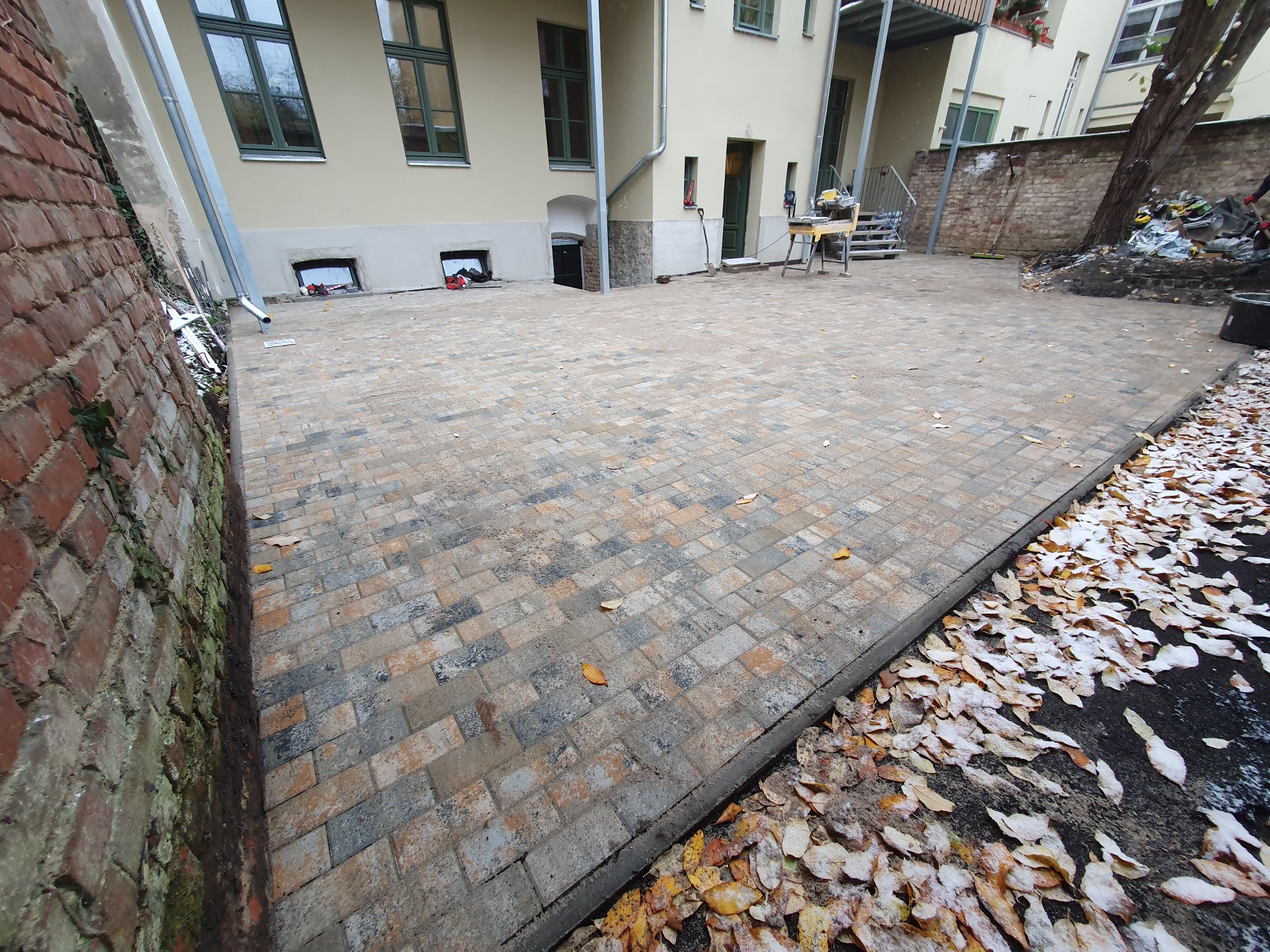 Nowo ułożona, wielobarwna kostka brukowa na posesji, z widoczną maszyną do cięcia kostki w tle i opadłymi liśćmi z lekkim śniegiem na obrzeżach.