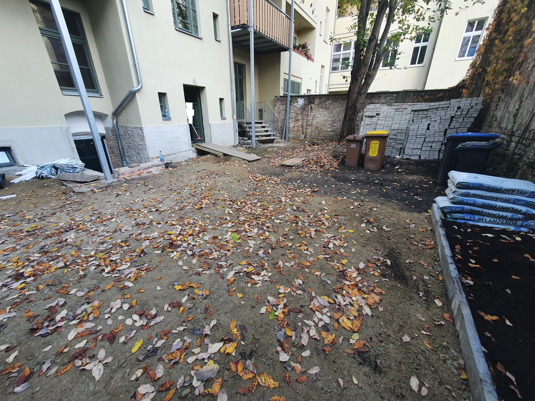 Przygotowywany teren pod przyszłą nawierzchnię, widoczne worki z ziemią, ułożone kostki, liście na ziemi, budynek w tle.