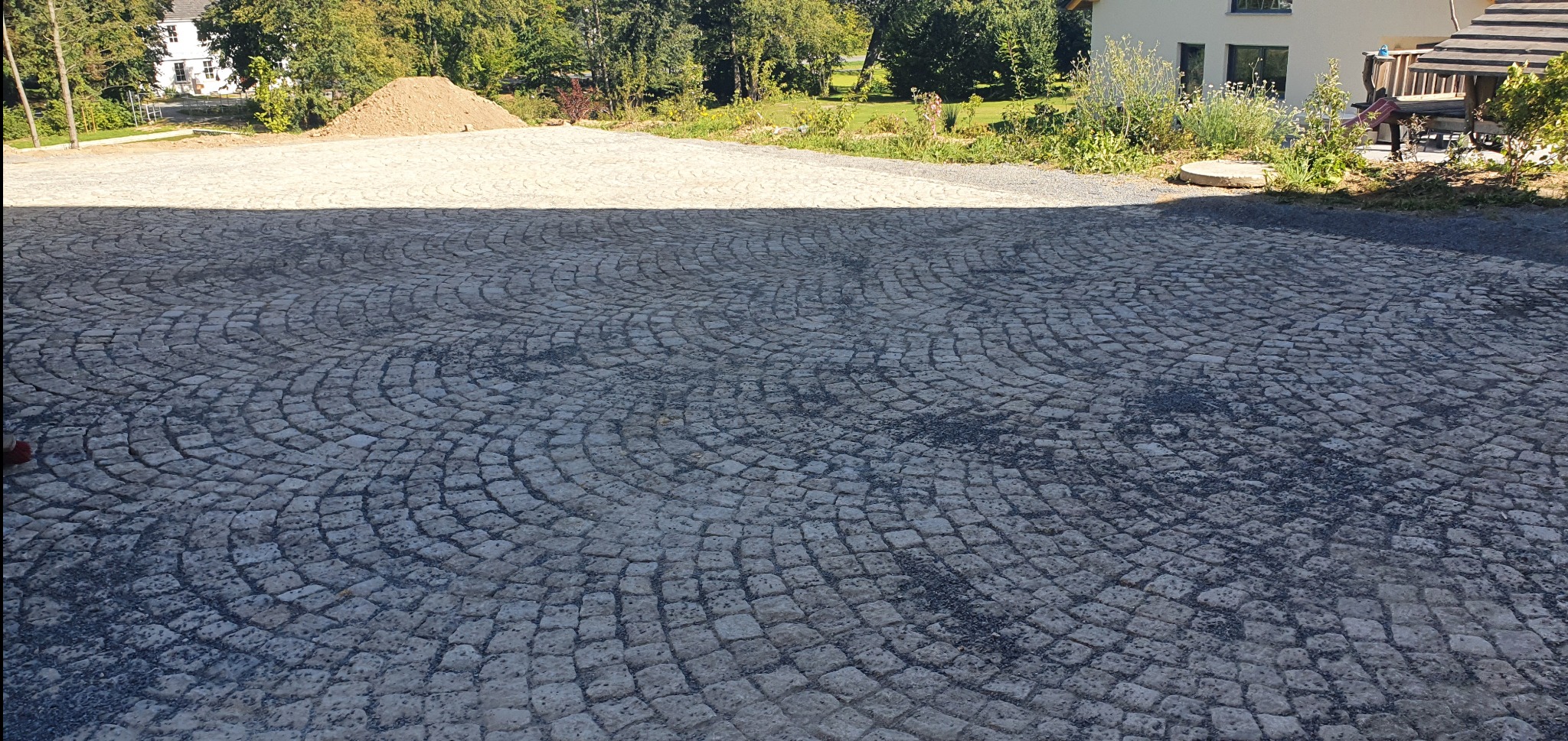 Ułożona nawierzchnia z kostki granitowej w kształcie łuków, z widocznym domem w tle i stertą ziemi.