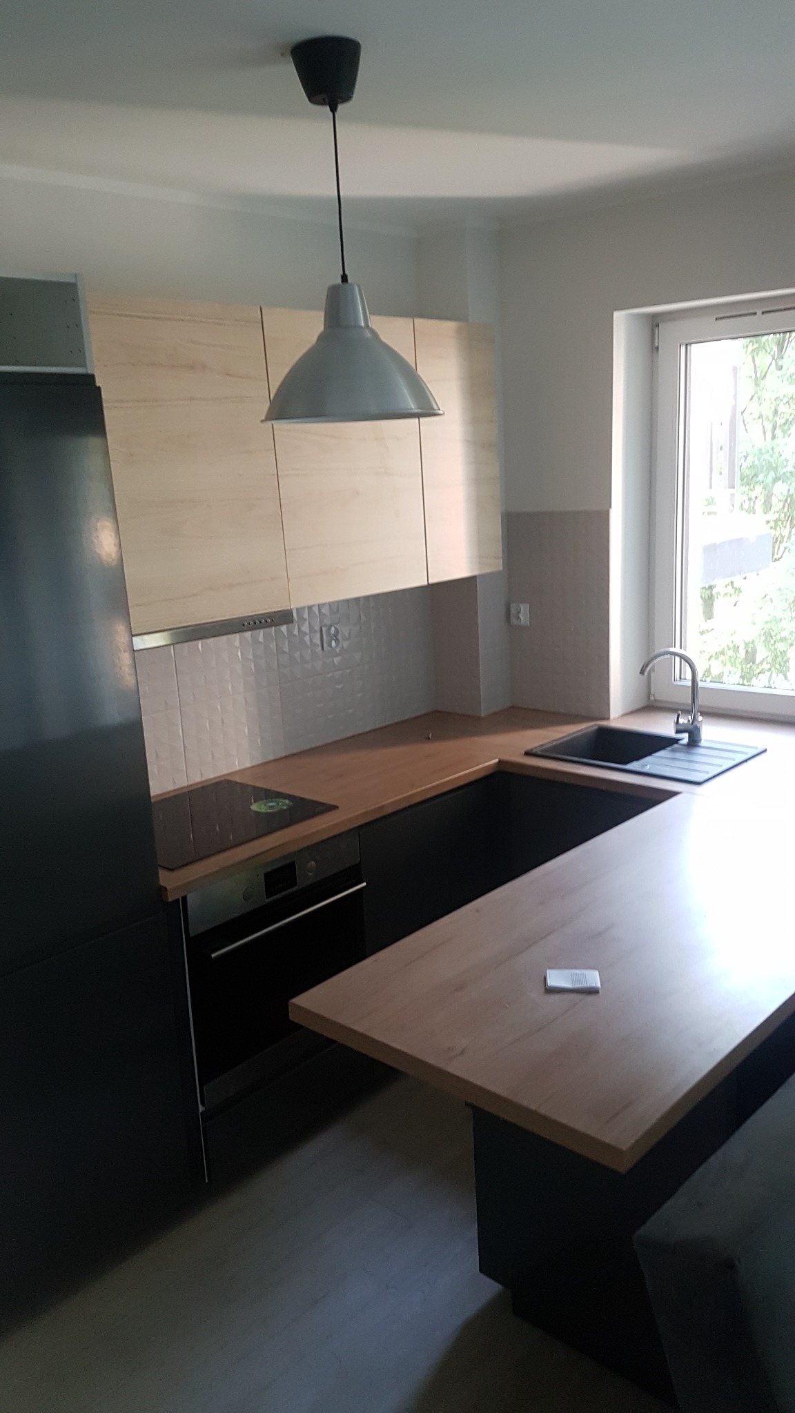 Nowoczesna kuchnia z jasnymi drewnianymi szafkami wiszącymi i czarnymi szafkami dolnymi, z blatem w kolorze drewna i płytą indukcyjną, oświetlona wiszącą lampą w kształcie dzwonu.