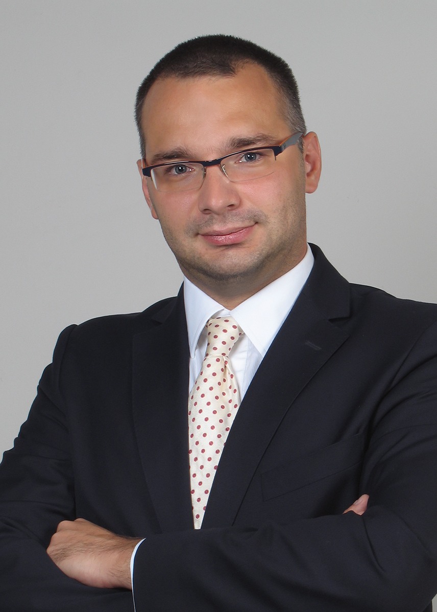 Portret mężczyzny w garniturze, z założonymi rękami, na jasnoszarym tle. Ma na sobie okulary w ciemnych oprawkach i krawat w jasne kropki.