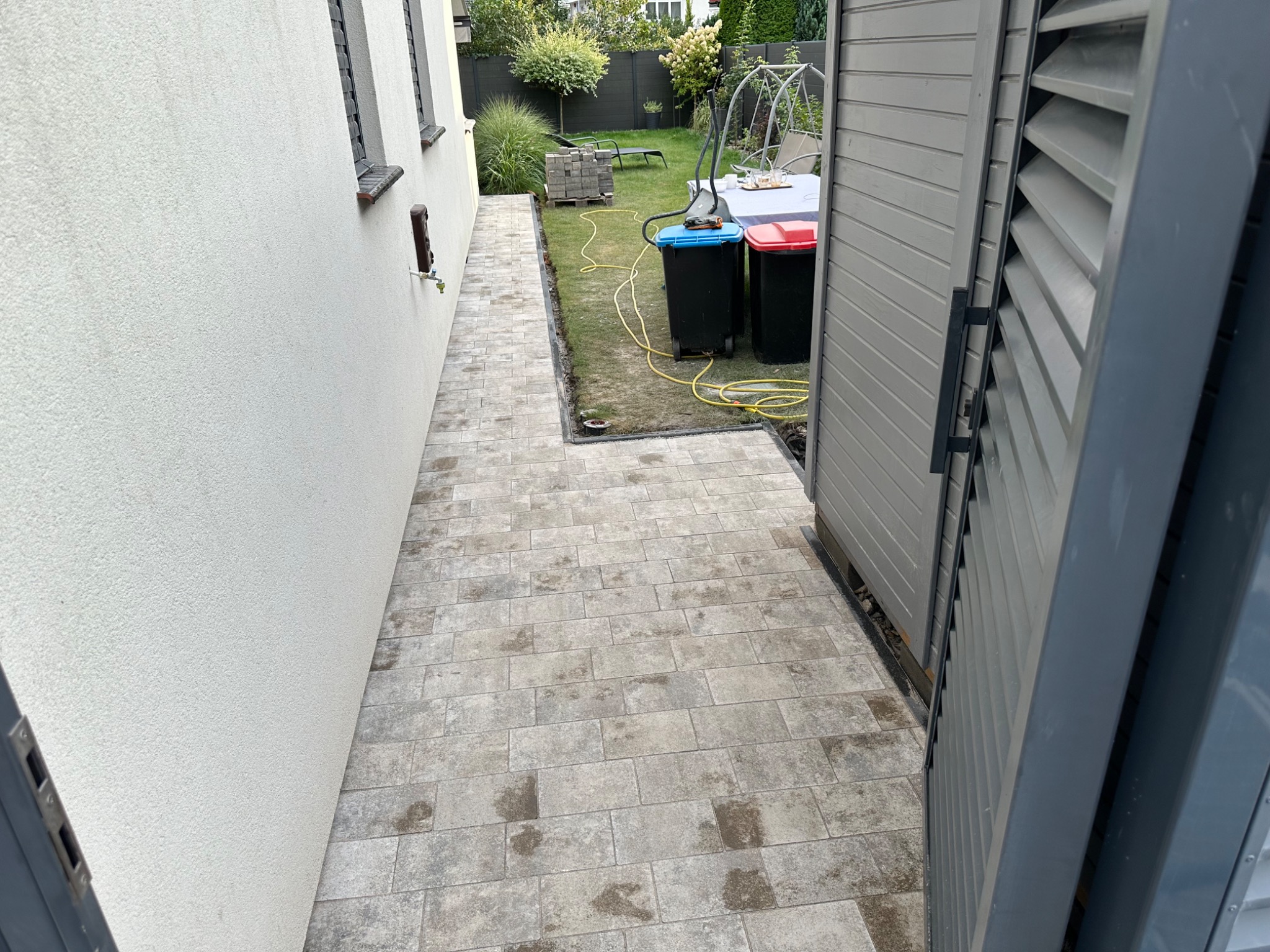 Ułożona ścieżka z kostki brukowej prowadząca wzdłuż ściany domu, widok z perspektywy otwartych drzwi szopy ogrodowej, na trawniku widoczne dwa kosze na śmieci i paleta kostki brukowej.