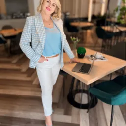 Uśmiechnięta blondynka w niebieskim żakiecie w pepitkę, białych spodniach i błękitnych szpilkach, opierająca się o jasny drewniany stół z laptopem, filiżanką kawy i dekoracją roślinną, w tle...