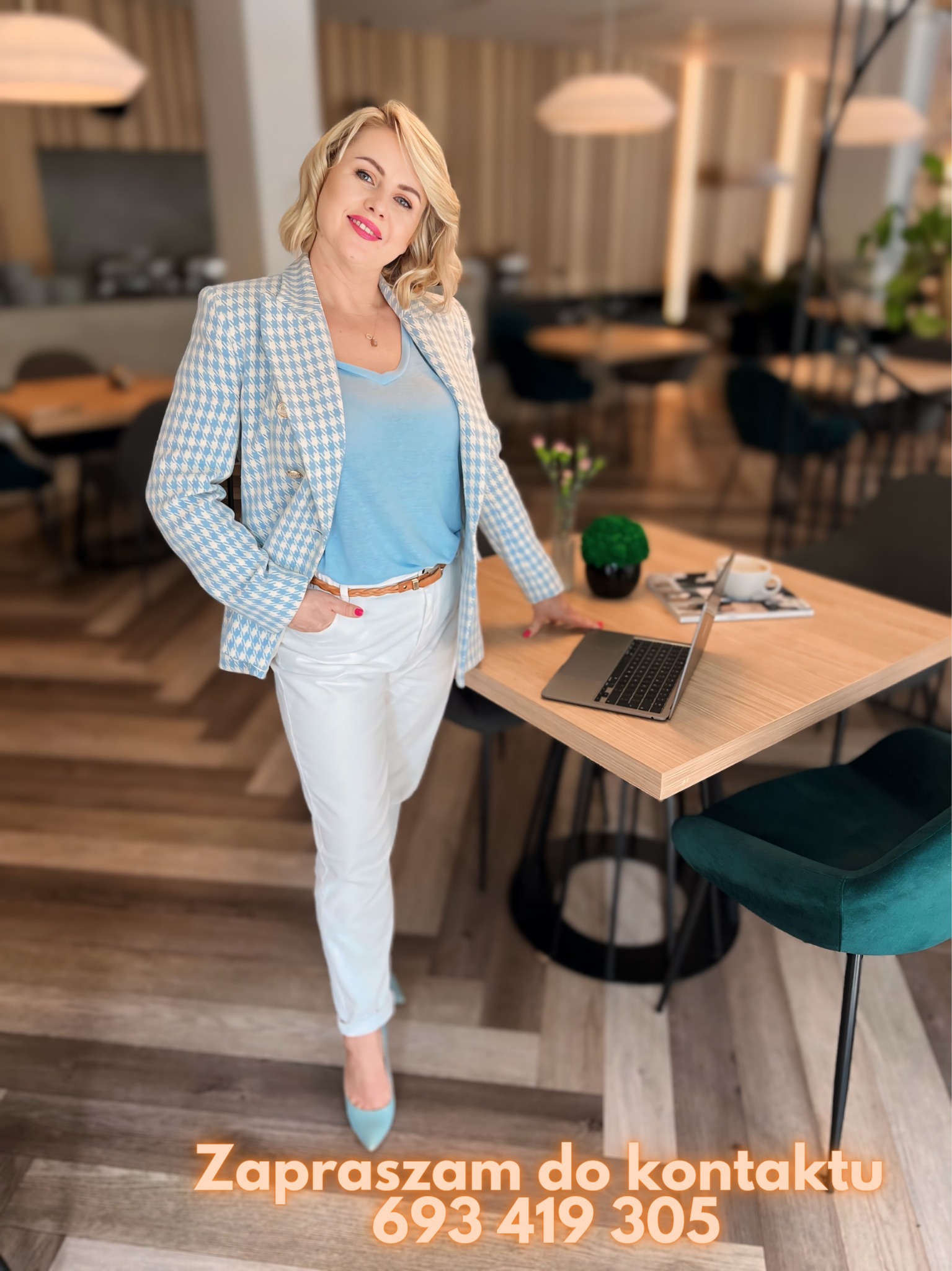 Uśmiechnięta blondynka w niebieskim żakiecie w pepitkę, białych spodniach i błękitnych szpilkach, opierająca się o jasny drewniany stół z laptopem, filiżanką kawy i dekoracją roślinną, w tle...