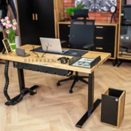 Drewniany blat biurka regulowanego elektrycznie z czarnymi nogami i organizerem kabli, laptop, biuro w tle