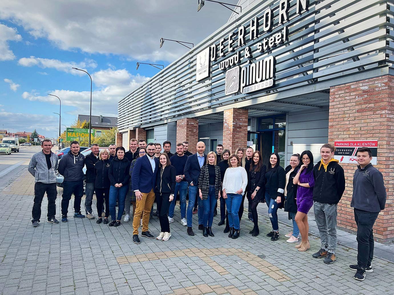 Zdjęcie grupowe pracowników firmy Deerhorn Wood & Steel przed budynkiem firmy z logo na elewacji i parkingiem dla klientów.
