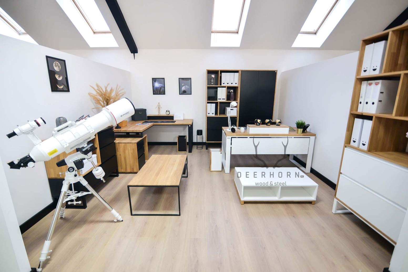 Jasne biuro z drewnianym blatem biurka narożnego, regałem z półkami, szafką z czarnymi drzwiami, stolikiem kawowym z logo Deerhorn oraz teleskopem na statywie.