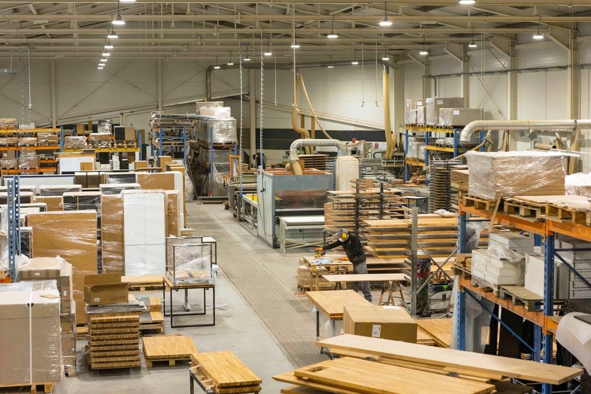 Wnętrze hali produkcyjnej z widocznymi regałami wypełnionymi elementami meblowymi, w tym blatami i frontami, niektóre zapakowane w folię, oraz pracownik szlifujący element drewniany przy pomocy...