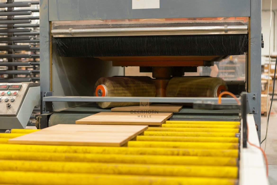 Obróbka drewnianych blatów w szlifierce przemysłowej; widoczne wałki szlifujące i posuwający się blat na żółtym transporterze rolkowym.