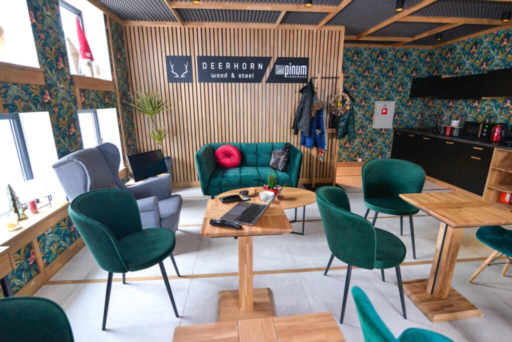 Nowoczesne wnętrze z drewnianymi blatami stołów, zielonymi krzesłami i sofą, drewnianą ścianą z logo firm Deerhorn Wood & Steel i Pinum Homewood, oraz kuchnią w tle.