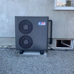 Zewnętrzna jednostka pompy ciepła Cooper&Hunter zainstalowana na betonowej podstawie, widok z poziomu gruntu na tle szarej ściany budynku z dwoma oknami.