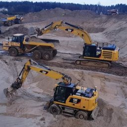 GRANAPOL SPÓŁKA Z OGRANICZONĄ ODPOWIEDZIALNOŚCIĄ - Żółta koparka CAT nabiera ziemię do ciężarówki BELL na placu budowy, w tle widoczne wzgórza piasku i inne maszyny.