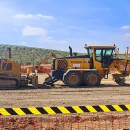 Żółty spychacz CAT i równiarka VOLVO G940 na wyrównanym terenie, z widoczną taśmą ostrzegawczą i drzewami w tle pod błękitnym niebem.