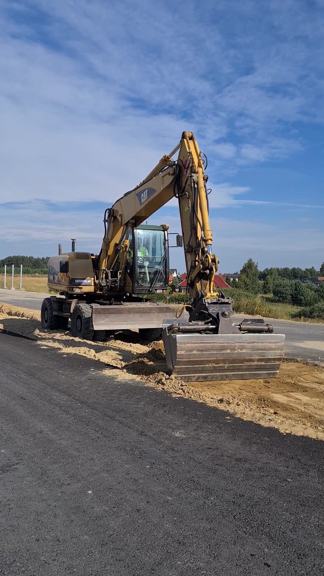 Żółta koparko-ładowarka Caterpillar wyrównuje pobocze drogi asfaltowej w słoneczny dzień.