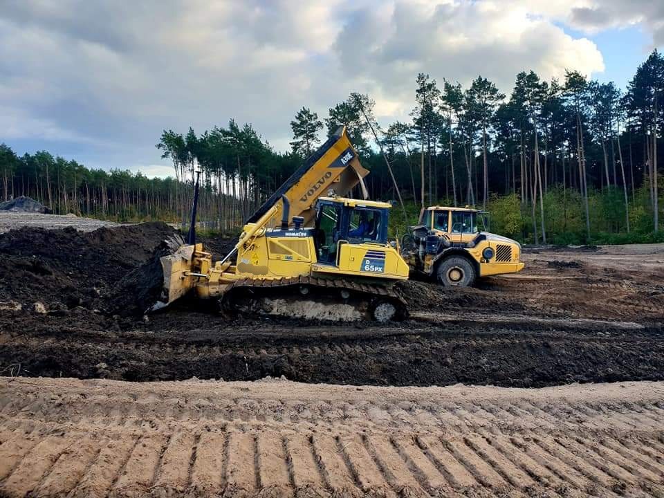 Żółty spychacz Komatsu D65PX w trakcie pracy na placu budowy, obok widoczna ładowarka Volvo, w tle las iglasty i pochmurne niebo.