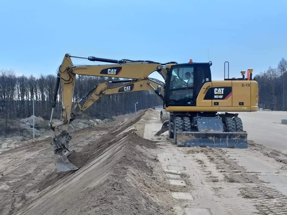 Żółta koparka kołowa CAT M316F wyrównuje teren piaszczystej skarpy na tle lasu i błękitnego nieba.
