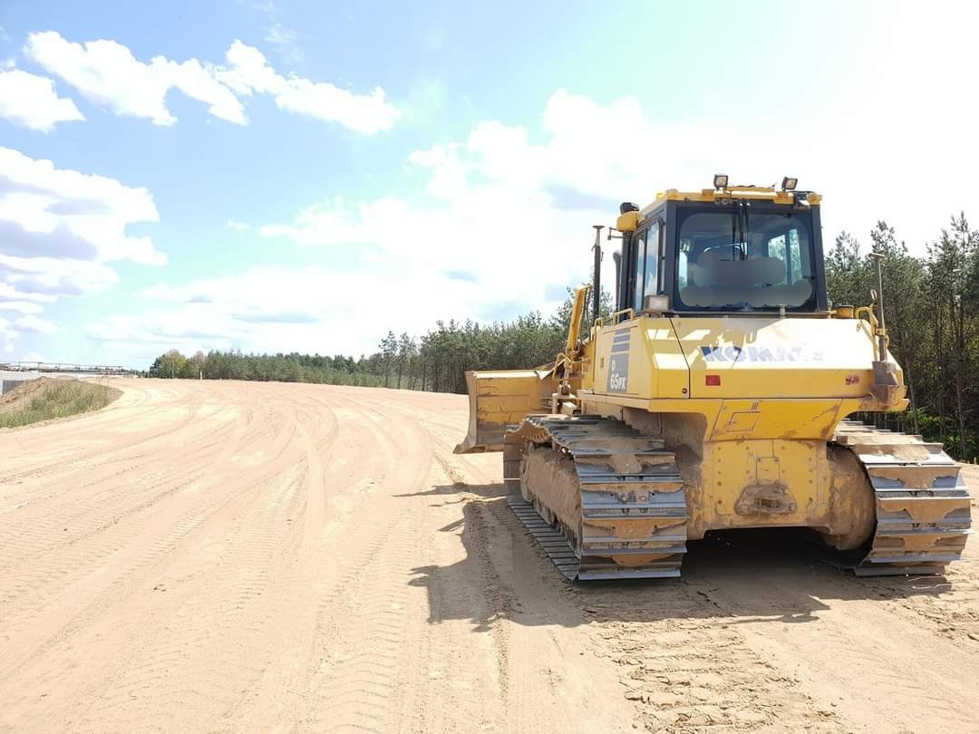 Żółty spychacz Komatsu 65PX na piaszczystym terenie budowy drogi, widoczne ślady gąsienic, w tle las i błękitne niebo z chmurami.