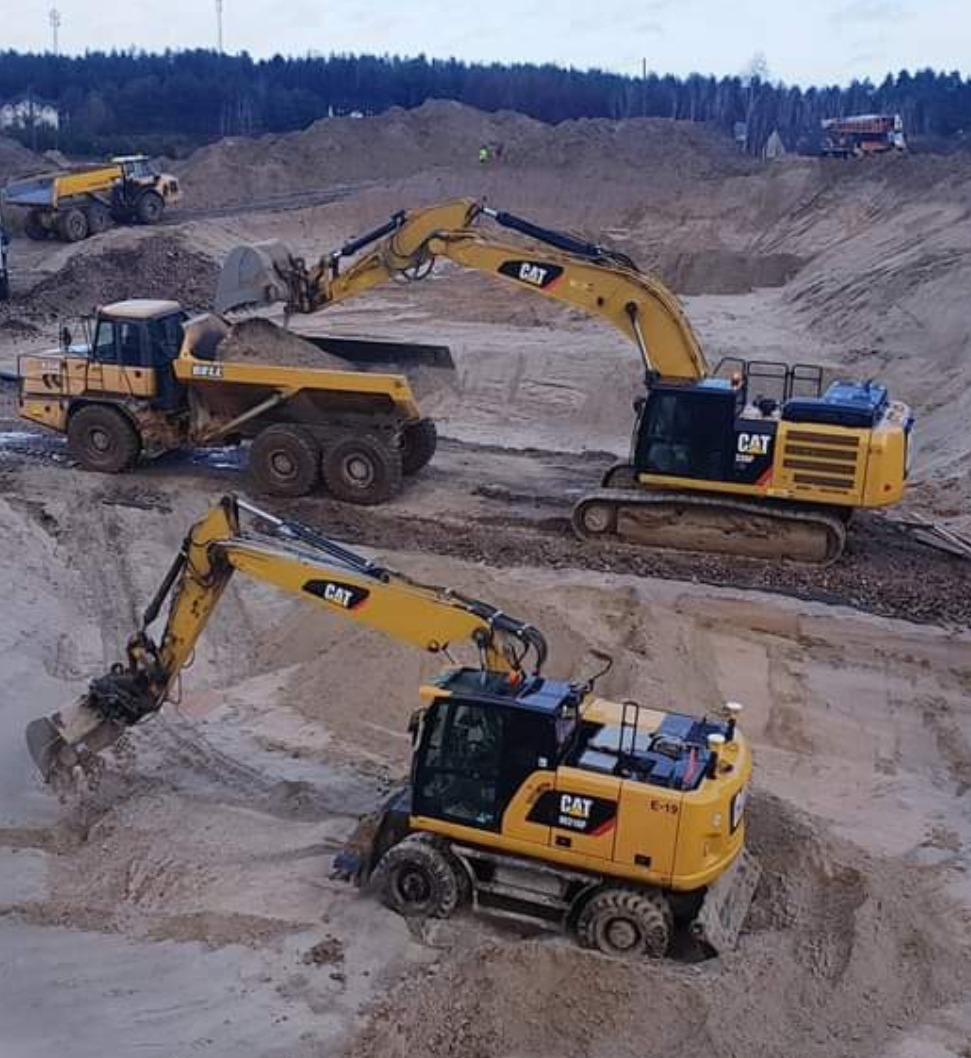 Żółta koparka CAT nabiera ziemię do ciężarówki BELL na placu budowy, w tle widoczne wzgórza piasku i inne maszyny.
